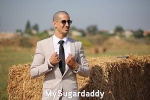 En la imagen vemos a un hombre vestido formalmente. Tiene un traje de color claro. Una sonrisa y unos lentes de sol oscuros. 