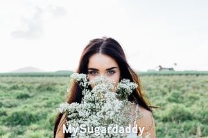 ¿Cómo debería ser una Sugar Baby?: En la imagen vemos a una mujer joven que se esconde detrás de un ramo de flores. Tiene el pelo suelto y una mirada profunda. Es muy atractiva. Se encuentra en un campo, pero el foco de la camera se dirige solamente a ella.  