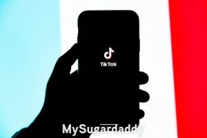 Sugar Baby TikTok: Applicación en teléfono celular Sugar Baby TikTok: En la imagen esta el logo de TikTok proyectado en un celular. Una silueta de una mano sostiene el celular y en el fondo hay colores llamativos.